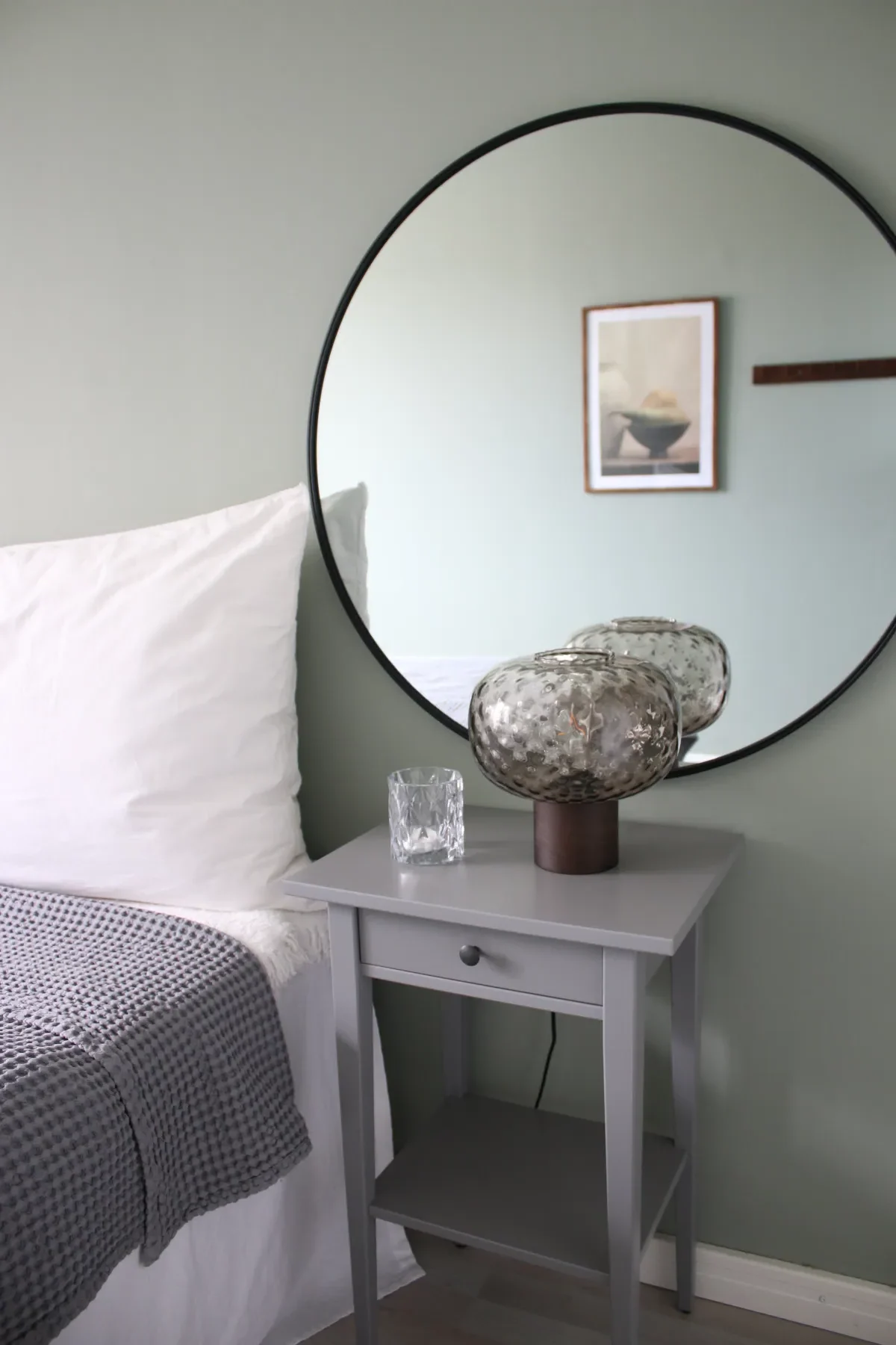 En minimalistisk sovrumsscen med ett grått nattduksbord med en texturerad glaslampa och en genomskinlig tumlare, bredvid en säng med vita kuddar och en grårutig filt. En stor rund spegel och inramad konst hänger ovanför.