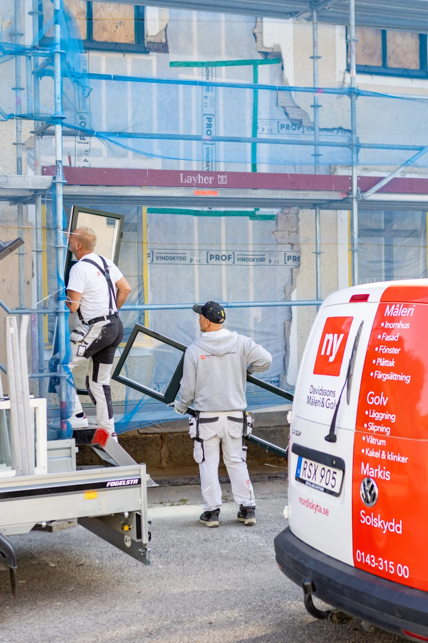 Två byggnadsarbetare står utanför en byggnad under renovering, varav en håller i en fönsterkarm. Byggnaden är täckt med ställningar och blått nät. En vit och röd arbetsbil med svensk text står parkerad i förgrunden.