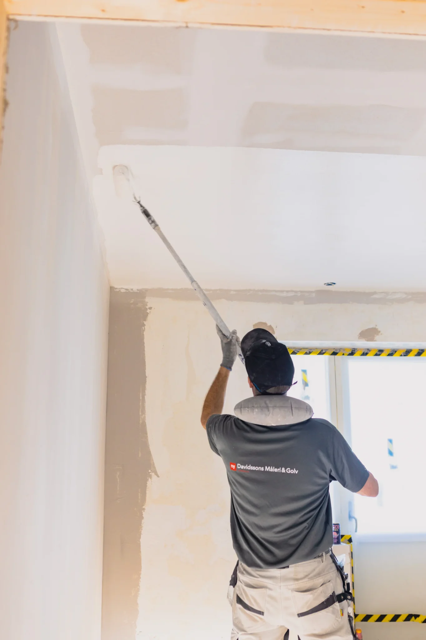 En person som bär en grå skjorta och vita byxor använder en rullborste med förlängningsstång för att måla ett vitt tak i ett rum under renovering.