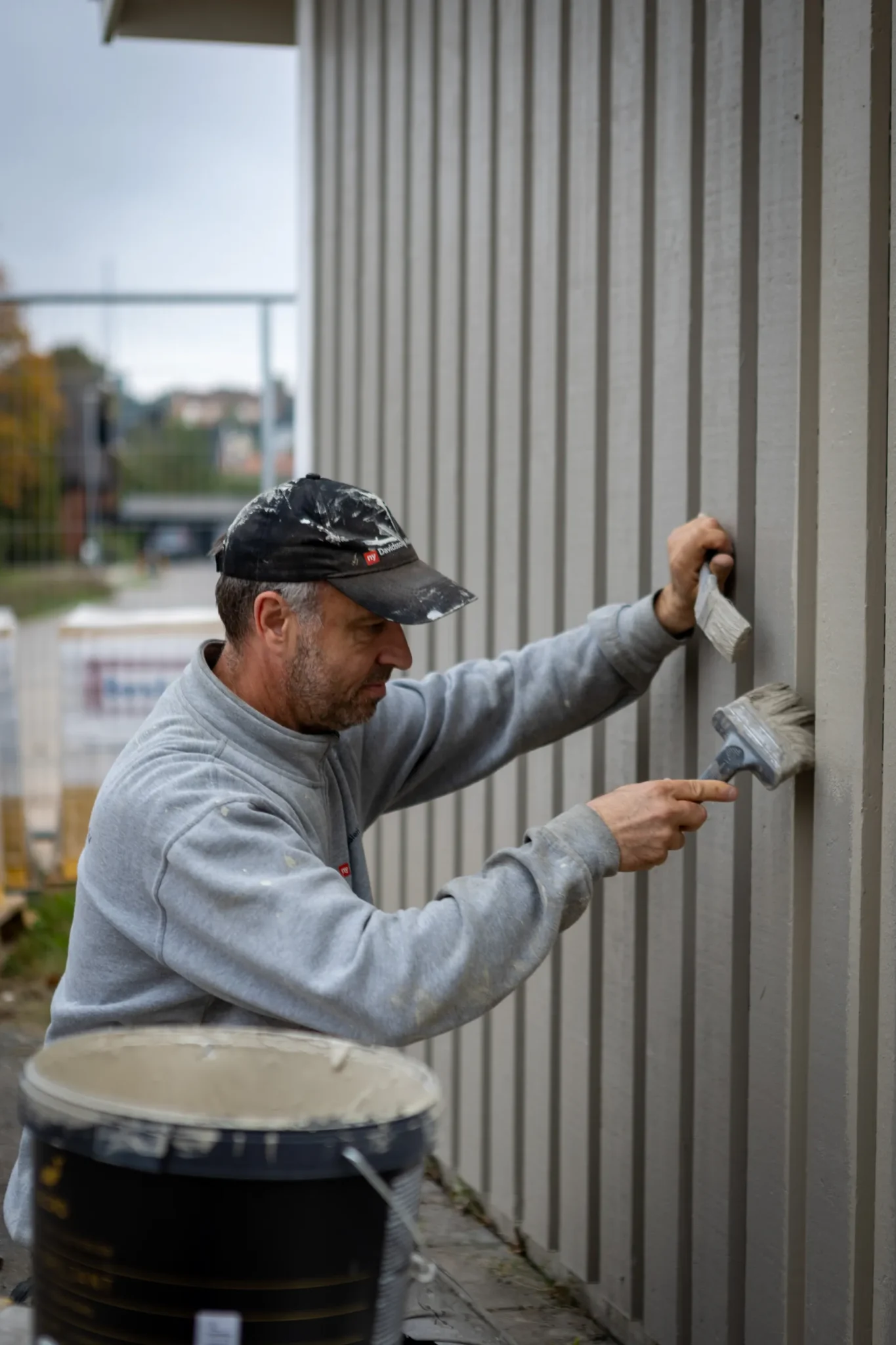 En man som bär keps och tröja målar utsidan av en träbyggnad med en pensel och håller fast i en panel som stöd; en hink med färg sitter i närheten.