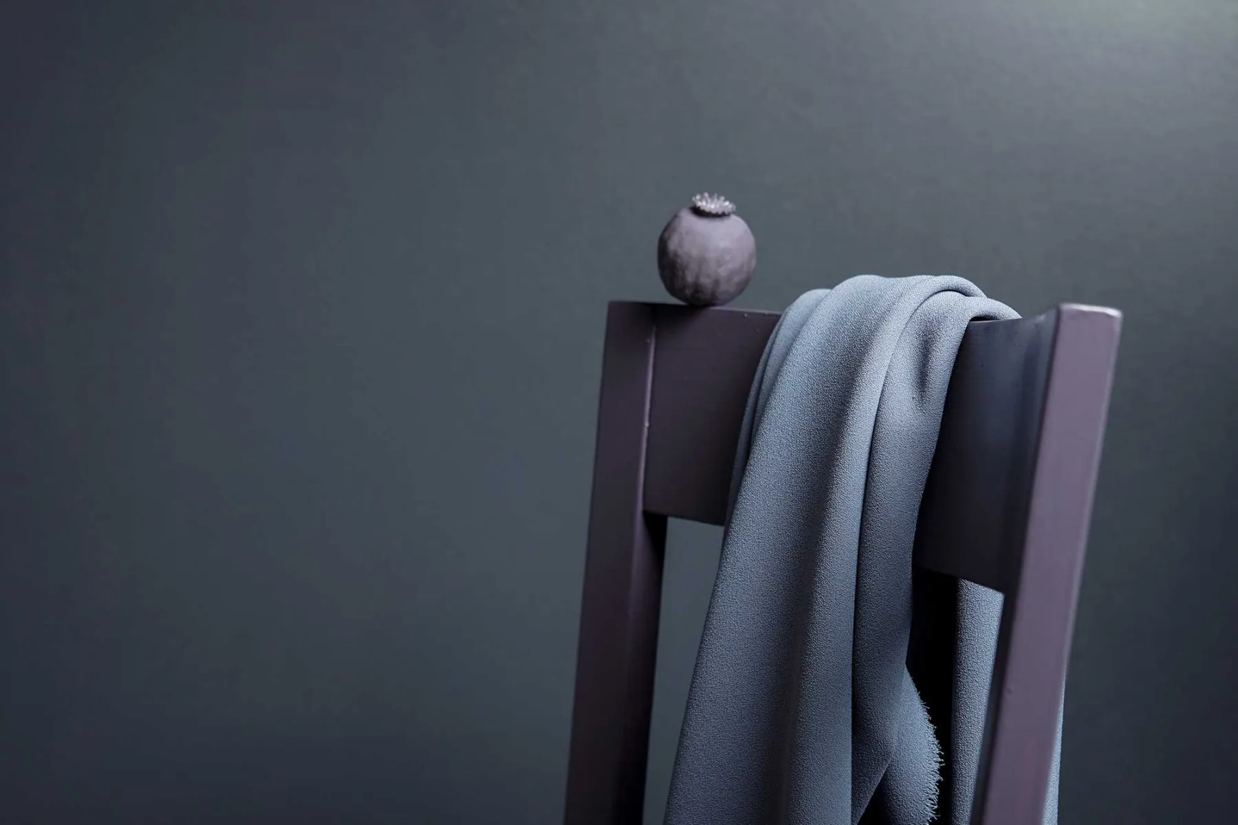 Ett grått tyg draperat över ryggen på en mörk trästol, med ett litet runt föremål, eventuellt en torkad fröskida, placerad ovanpå stolen mot en vanlig mörk bakgrund.