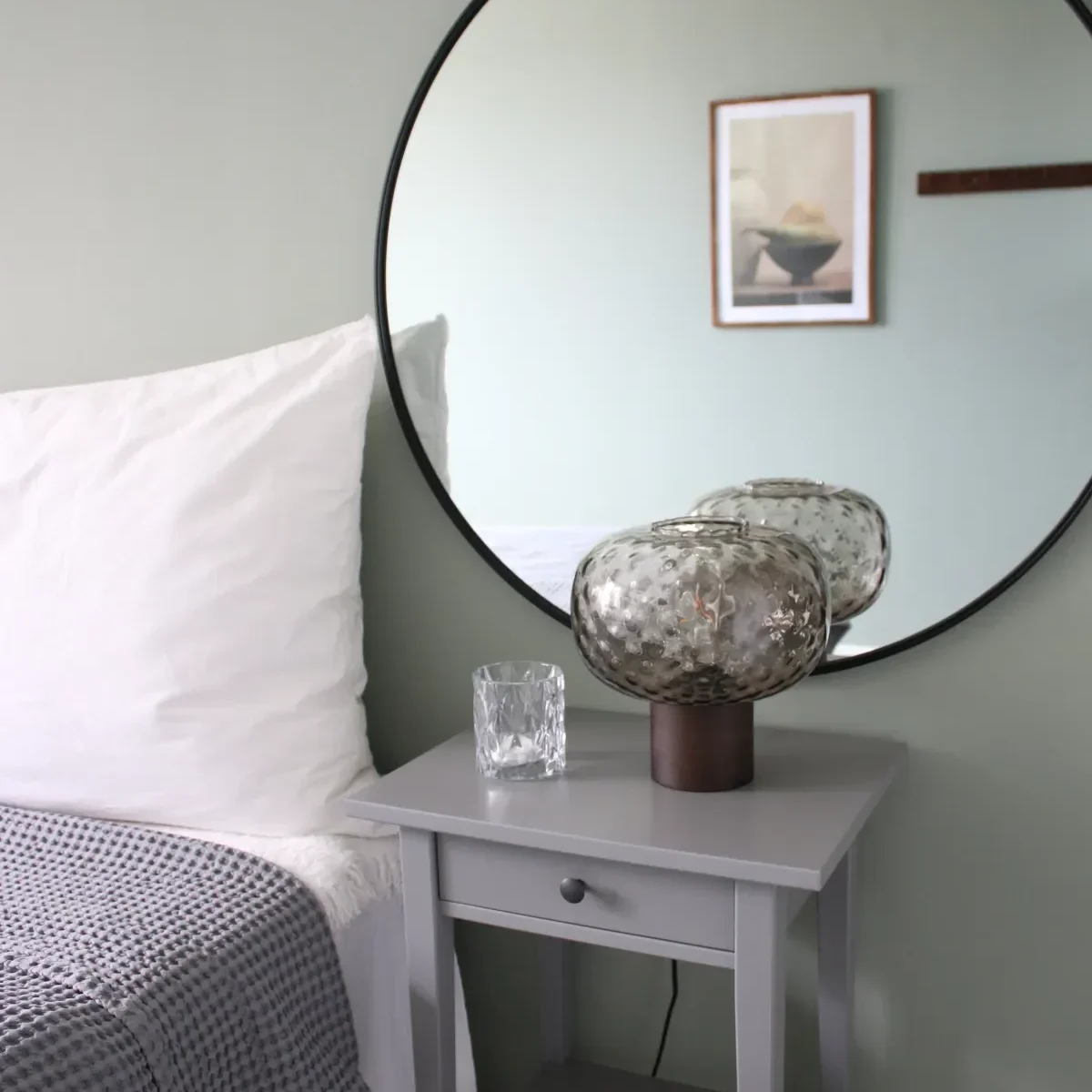 En minimalistisk sovrumsscen med ett grått nattduksbord med en texturerad glaslampa och en genomskinlig tumlare, bredvid en säng med vita kuddar och en grårutig filt. En stor rund spegel och inramad konst hänger ovanför.