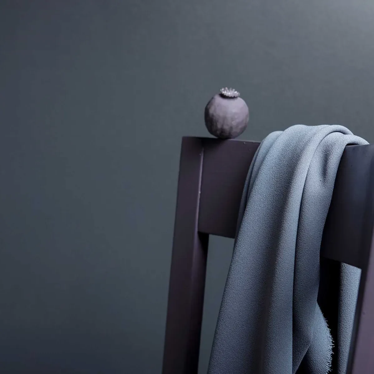 Ett grått tyg draperat över ryggen på en mörk trästol, med ett litet runt föremål, eventuellt en torkad fröskida, placerad ovanpå stolen mot en vanlig mörk bakgrund.